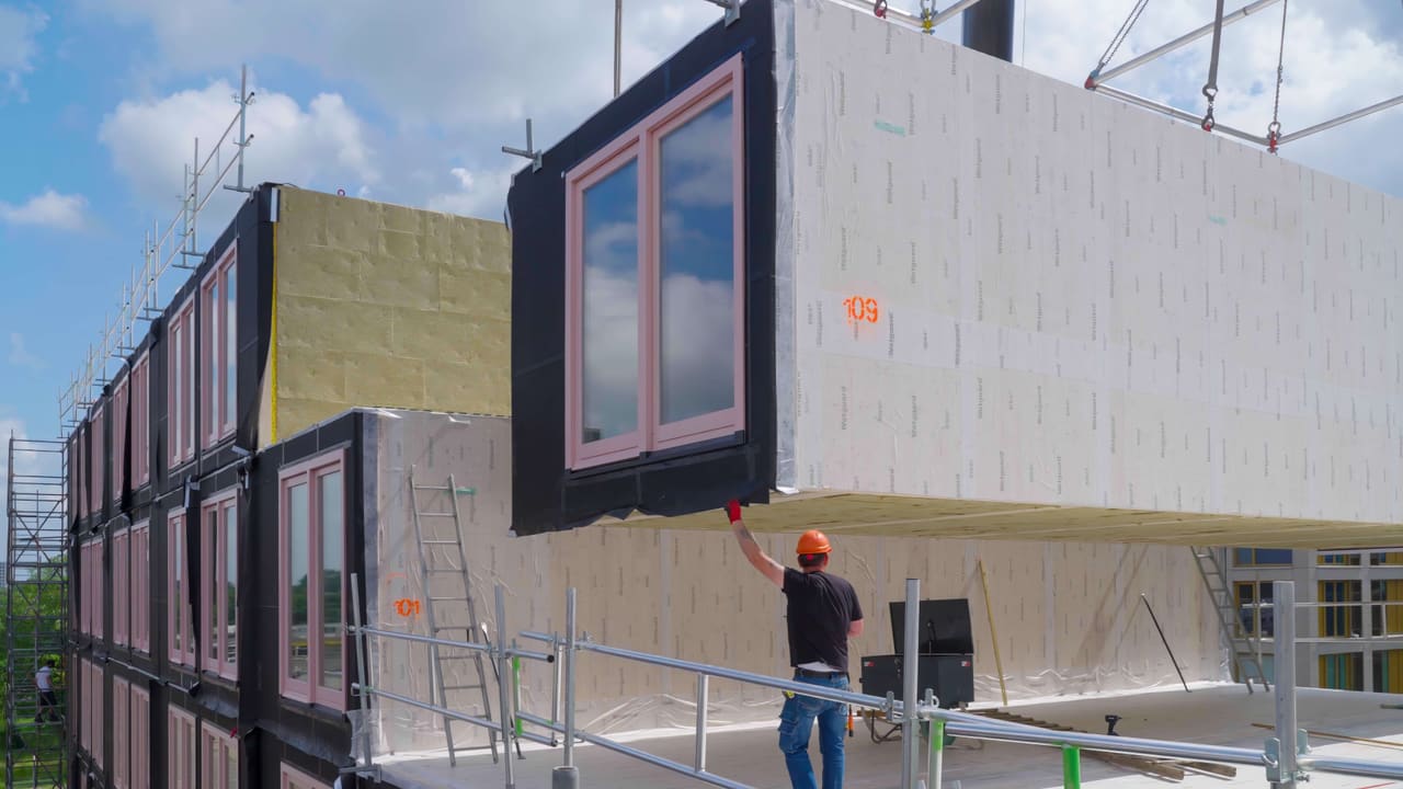 A prefabricated timber module with pink window frames is lowered into place at Poppies